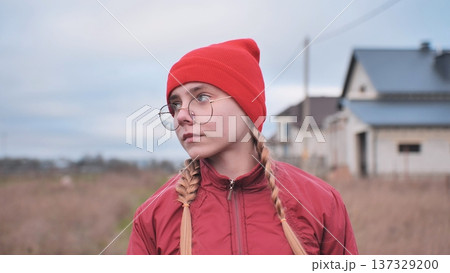 Portrait of a pensive teenage girl wearing a red beanie and glasses, standing in a field and looking away 137329200