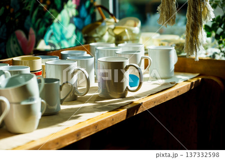 Row of white mugs in warm sunlight on rustic shelf. Coffee shop interior, morning routine and small business atmosphere. Row of white mugs in warm sunlight on rustic shelf. Coffee shop interior, morning routine and small business atmosphere. 137332598