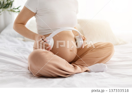 Pregnant woman placing headphones on belly while sitting on bed at home Pregnant woman placing headphones on belly while sitting on bed at home 137333518