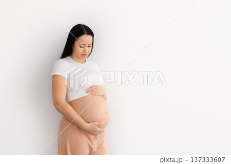 Pregnant beautiful woman standing against white wall background, gently holding belly Pregnant beautiful woman standing against white wall background, gently holding belly 137333607
