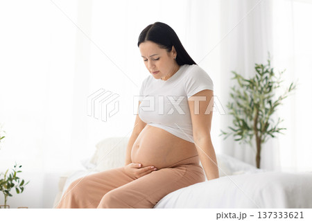 Young pregnant woman sitting on bed at home holding belly and frowning Young pregnant woman sitting on bed at home holding belly and frowning 137333621