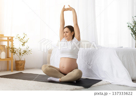 Young pregnant woman sitting on yoga mat at home stretching arms upward Young pregnant woman sitting on yoga mat at home stretching arms upward 137333622