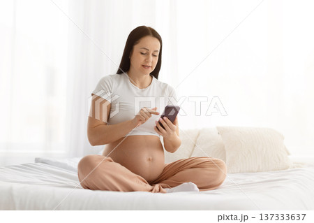 Young pregnant woman using smartphone while sitting on bed at home Young pregnant woman using smartphone while sitting on bed at home 137333637
