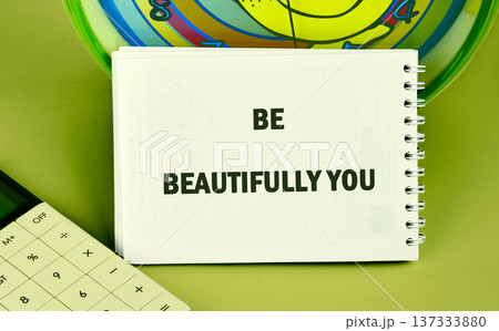 A pristine white notebook displays the powerful words BE BEAUTIFULLY YOU on a vibrant green desktop a partial calculator and colorful disk nearby 137333880
