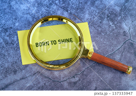 A golden framed magnifying glass magnifies the powerful message Born to Shine on a vibrant yellow note, thoughtfully placed on a dark blue marbled surface 137333947