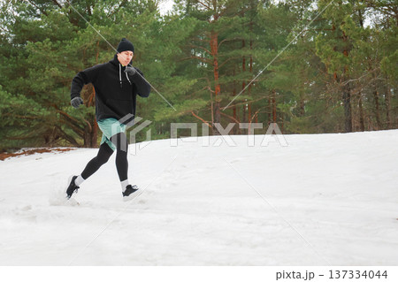 Young man running in a snowy pine forest. Winter outdoor fitness workout. Male runner exercising in cold weather wearing a black hoodie and beanie. High quality photo Young man running in a snowy pine forest. Winter outdoor fitness workout. Male runner exercising in cold weather wearing a black hoodie and beanie. High quality photo 137334044