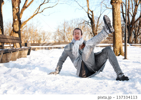 Young man falling on slippery snow in a winter park. Guy looking in pain after an accident outdoors. Concept of injury, insurance and safety during cold season. High quality photo Young man falling on slippery snow in a winter park. Guy looking in pain after an accident outdoors. Concept of injury, insurance and safety during cold season. High quality photo 137334113