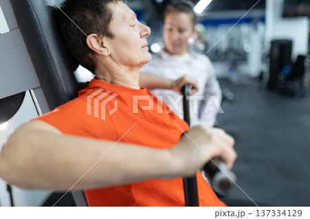 Senior woman exercising on gym machine with trainer Senior woman exercising on gym machine with trainer 137334129