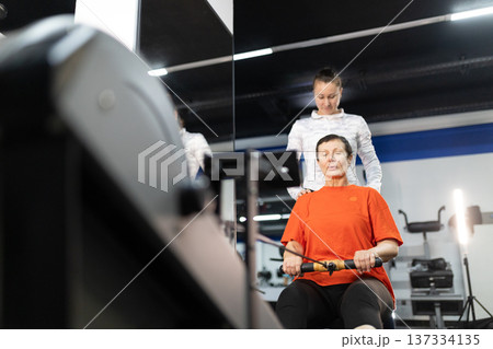 Senior woman training on rowing machine with personal trainer Senior woman training on rowing machine with personal trainer 137334135