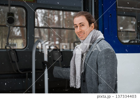 Smiling young man standing in the open doorway of a tram during a snowfall. Happy commuter in a grey coat and scarf traveling by public transport in winter. High quality photo Smiling young man standing in the open doorway of a tram during a snowfall. Happy commuter in a grey coat and scarf traveling by public transport in winter. High quality photo 137334403