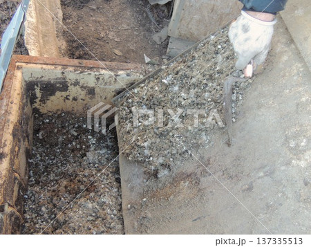A farm worker wearing gloves cleans an iron tray of bird droppings with a spatula, and during the cleaning process, transfers organic waste from caged quails from the tray into a large iron container. 137335513