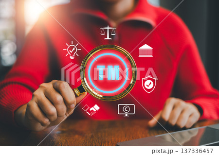 Person in red sweater holding magnifying glass with trademark symbol, surrounded by business icons, emphasizing intellectual property. Person in red sweater holding magnifying glass with trademark symbol, surrounded by business icons, emphasizing intellectual property. 137336457