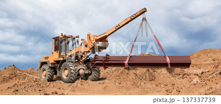 At a construction site, a knee loader lifts a steel beam. The area is littered with mounds of earth, and the sky is cloudy. Heavy and long loads are being transported 137337739