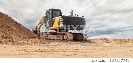 An excavator works on a construction site moving earth and digging into sandy soil while clouds cover the sky in the background during the day 137337743