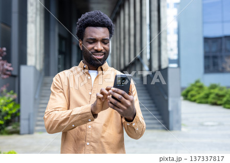 Young adult man smiling while using a smartphone for communication and social media, engaging with digital technology in a contemporary urban environment Young adult man smiling while using a smartphone for communication and social media, engaging with digital technology in a contemporary urban environment 137337817