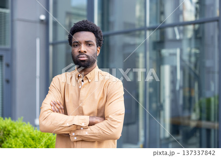 Young man with a serious expression and crossed arms stands confidently in front of a modern glass building, portraying professionalism, determination, and competence in an urban setting 137337842