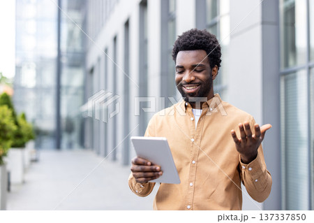Young man smiling and gesturing while having a video conference on his digital tablet, standing outdoors in a modern urban setting, engaging in remote communication 137337850