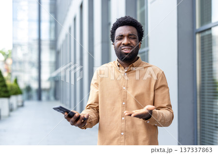 Young adult man shrugging with a confused expression, holding a mobile phone and gesturing with his open hand, depicting themes of confusion, problems, question, and difficulty in an urban setting Young adult man shrugging with a confused expression, holding a mobile phone and gesturing with his open hand, depicting themes of confusion, problems, question, and difficulty in an urban setting 137337863