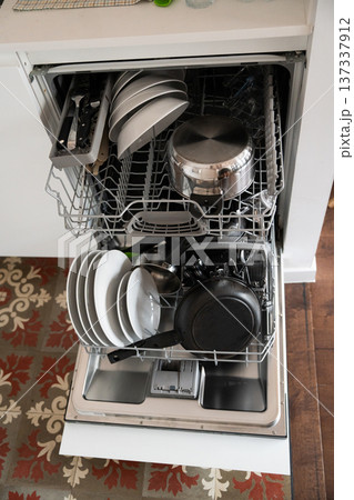 Fully loaded dishwasher featuring various dirty kitchen items like plates, bowls, pots, pans, and silverware, representing daily household chores and modern convenience in a kitchen 137337912