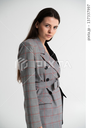 Young brunette woman posing in a studio, wearing an elegant plaid blazer dress with a belted waist, showcasing modern fashion and professional style 137337997