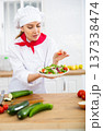 Smiling female chef in white uniform preparing vegetable salad in private kitchen 137338474