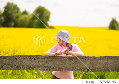 Young woman wearing a bucket hat leaning on an old wooden fence with a blooming rapeseed field in the background 137342143