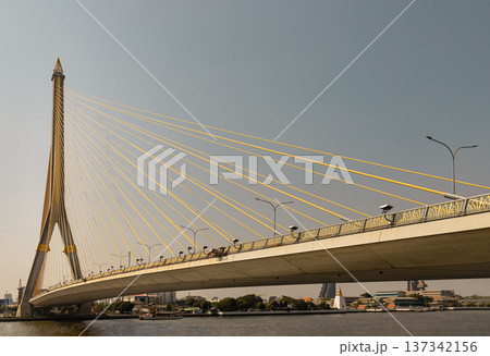 An elegant feat of engineering, the Rama VIII Bridge is anchored by a singular pylon an inverted 'Y' reaching toward the sky with a golden, flame-tipped spire. Golden-yellow cables extend from its An elegant feat of engineering, the Rama VIII Bridge is anchored by a singular pylon an inverted 'Y' reaching toward the sky with a golden, flame-tipped spire. Golden-yellow cables extend from its 137342156