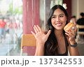 Young woman sitting in a cafe holding a glazed donut while waving her hand in greeting 137342553