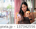Young Woman Looking at Smartphone While Holding Donut in Cafe 137342556