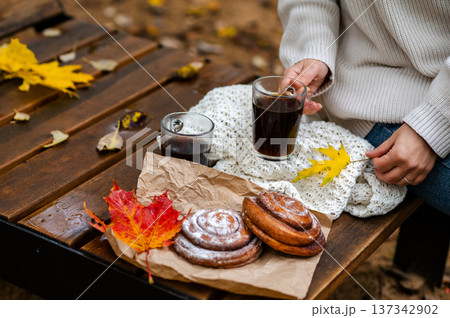Partial view of woman holding cup of coffee on wooden bench with cinnamon rolls and fall leave 137342902