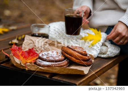 Partial view of woman holding cup of coffee on wooden bench with cinnamon rolls and fall leave 137342903