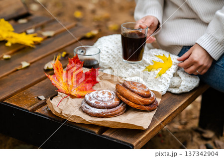 Partial view of woman holding cup of coffee on wooden bench with cinnamon rolls and fall leave 137342904