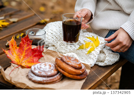 Partial view of woman holding glass cup of coffee beside cinnamon rolls and maple leaves on wooden bench 137342905