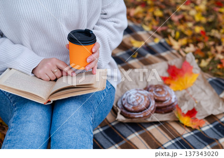 Partial view of woman sitting on blanket with book on knees and takeaway coffee during cozy autumn picnic in the park Partial view of woman sitting on blanket with book on knees and takeaway coffee during cozy autumn picnic in the park 137343020