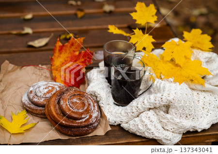 Cozy autumn picnic scene with two cups of coffee, cinnamon rolls and colorful maple leaves on a wooden bench wrapped in a white knitted blanket Cozy autumn picnic scene with two cups of coffee, cinnamon rolls and colorful maple leaves on a wooden bench wrapped in a white knitted blanket 137343041