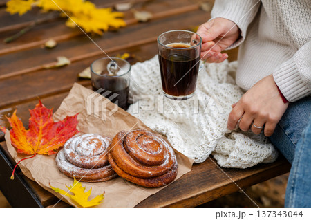 Cozy autumn picnic with a woman in a knitted sweater holding a glass cup of coffee next to cinnamon rolls and colorful maple leaves on a wooden bench 137343044