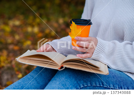 Partial view of woman holding takeaway coffee cup and open book on knees with autumn leaf outdoors in fall park Partial view of woman holding takeaway coffee cup and open book on knees with autumn leaf outdoors in fall park 137343088