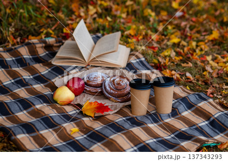 Cozy autumn picnic scene with plaid, two paper cups of hot drinks, pastries, apples and a book Cozy autumn picnic scene with plaid, two paper cups of hot drinks, pastries, apples and a book 137343293