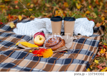 Cozy autumn picnic scene with plaid, knitted sweater, two paper cups of hot drinks, pastries, apples and falling maple leaves 137343302