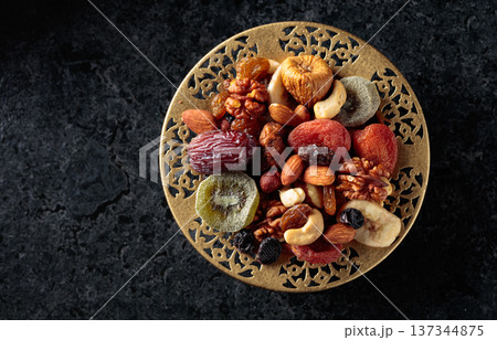 Dried tropical fruits, nuts, and raisins in a golden bowl on black stone table. 137344875