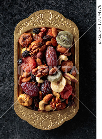 Dried tropical fruits, nuts, and raisins in a golden bowl on black stone table. 137344876