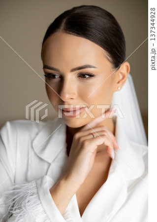Beautiful bride wearing white silk pajamas, touching her chin and closing her eyes 137344928