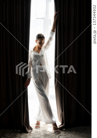 Bride opening curtains in white silk pajamas on wedding day Bride opening curtains in white silk pajamas on wedding day 137344932