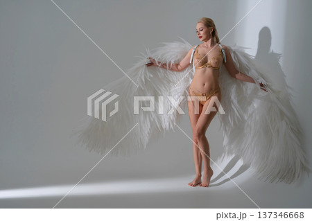 Elegant woman in gold lingerie posing with large white angel wings in a minimalist studio. 137346668