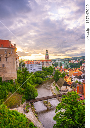 Travelers explore Cesky Krumlov Castle in Czechia on a summer day. The historic site overlooks the town, with green trees and a river in the foreground. Clouds fill the sky. 137347049