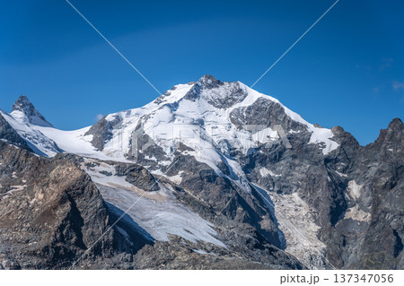 Piz Bernina showcases its stunning peaks against a backdrop of clear blue skies. The alpine landscape features glaciers and rugged mountains in Graubunden, Switzerland, creating a breathtaking scene. 137347056
