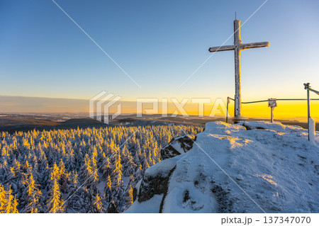 A cross stands on the summit rock of Jizera Mountain. Snow covers the ground and trees. Sunset casts warm light over the landscape, creating a striking contrast with the winter scene. A cross stands on the summit rock of Jizera Mountain. Snow covers the ground and trees. Sunset casts warm light over the landscape, creating a striking contrast with the winter scene. 137347070