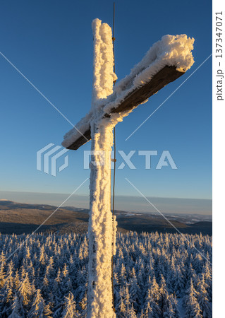 A cross stands tall on the summit rock formation of Jizera Mountain. Winter blankets the surrounding trees in snow. The sky is clear, showing a bright blue above the landscape. A cross stands tall on the summit rock formation of Jizera Mountain. Winter blankets the surrounding trees in snow. The sky is clear, showing a bright blue above the landscape. 137347071