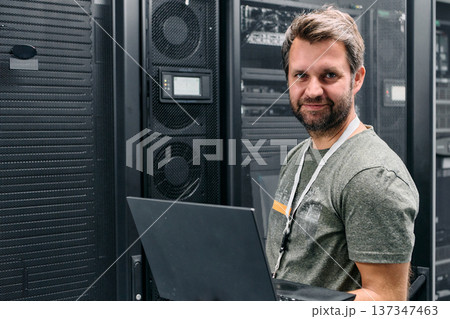 Smiling IT technician working on laptop in a server room surrounded by racks and equipment Smiling IT technician working on laptop in a server room surrounded by racks and equipment 137347463