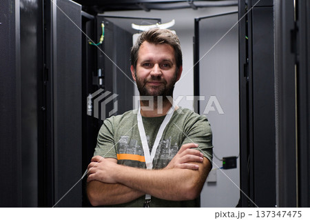 Confident IT technician standing with arms crossed between server racks in a modern data center 137347475